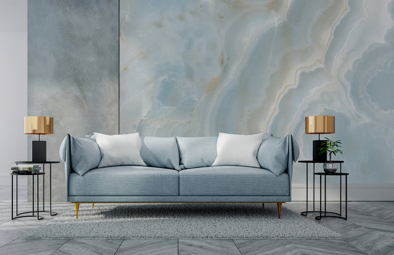 May include: A light blue sofa with white pillows sits in front of a wall with a blue and white marble pattern. Two black side tables with gold lamps are on either side of the sofa. A gray rug is on the floor.