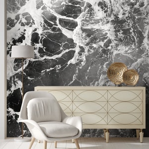 May include: A modern living room featuring a black and white abstract wallpaper resembling ocean waves. The room includes a white armchair, a cream-colored cabinet with gold accents, a floor lamp, and decorative gold plates.