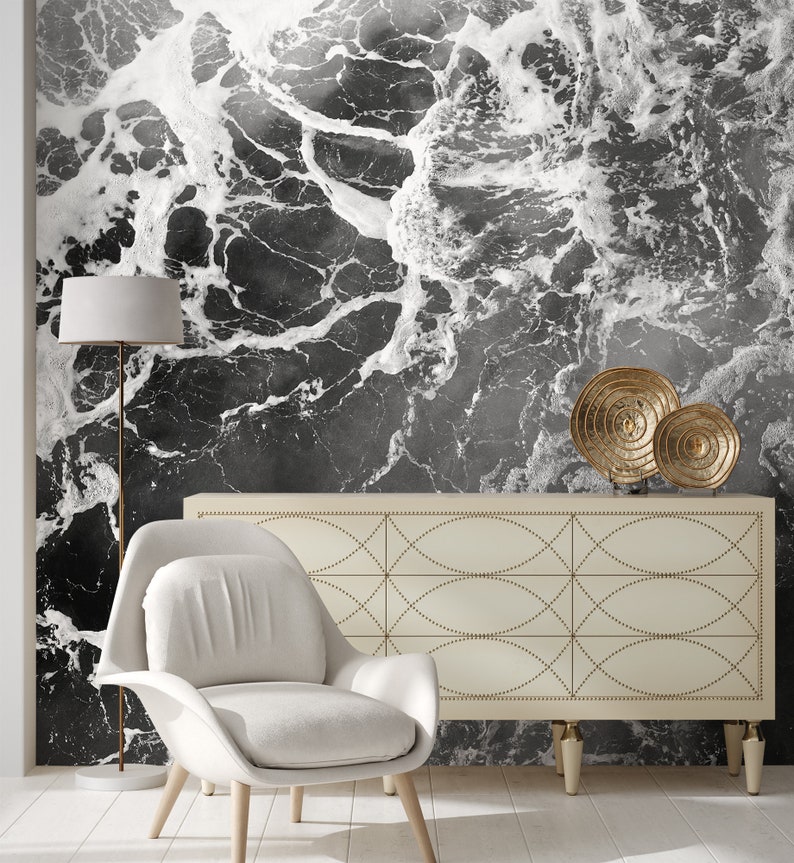May include: A modern living room featuring a black and white abstract wallpaper resembling ocean waves. The room includes a white armchair, a cream-colored cabinet with gold accents, a floor lamp, and decorative gold plates.