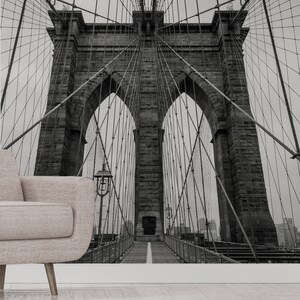 May include: A black and white photograph of the Brooklyn Bridge in New York City. The bridge is a suspension bridge with two towers and a walkway in the center. The photo is taken from below the bridge, looking up at the towers.