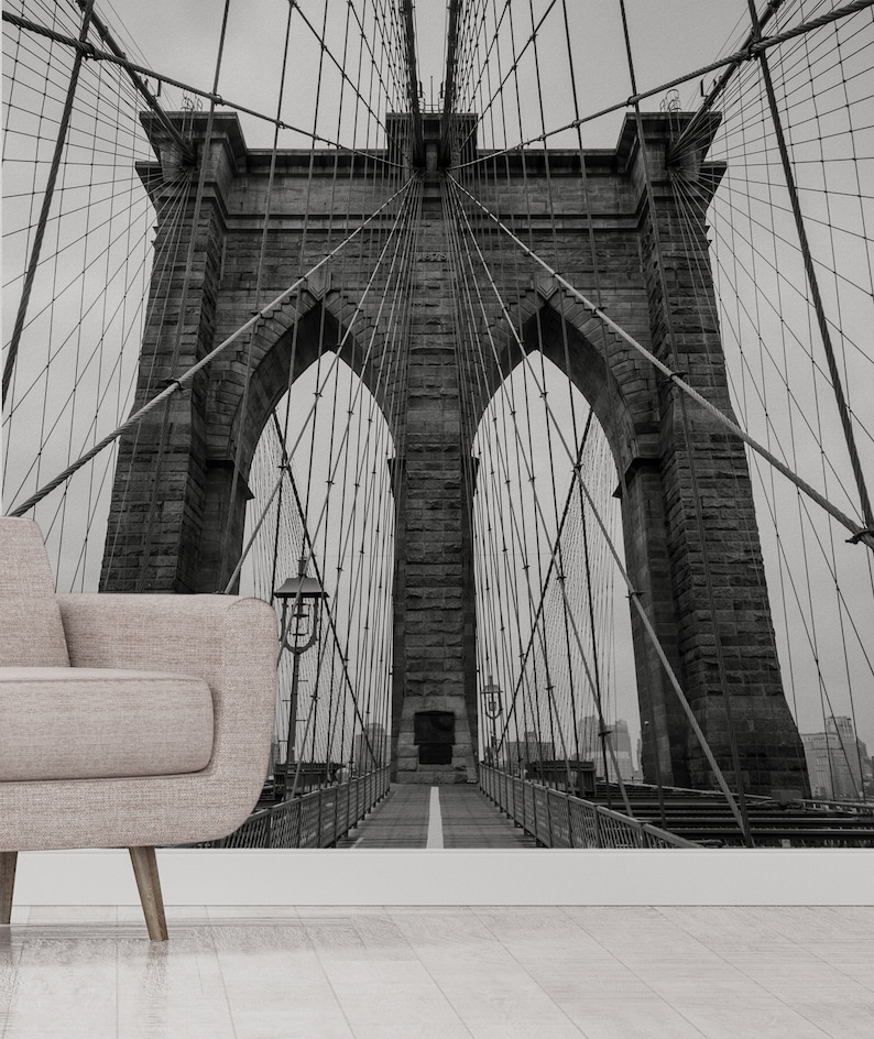 May include: A black and white photograph of the Brooklyn Bridge in New York City. The bridge is a suspension bridge with two towers and a walkway in the center. The photo is taken from below the bridge, looking up at the towers.