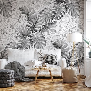 May include: A white sofa with a black and white throw blanket and a black and white patterned pillow sits in front of a wall with a black and white tropical leaf wallpaper. A small table with a vase of dried branches and a lamp with a white shade are in the room.