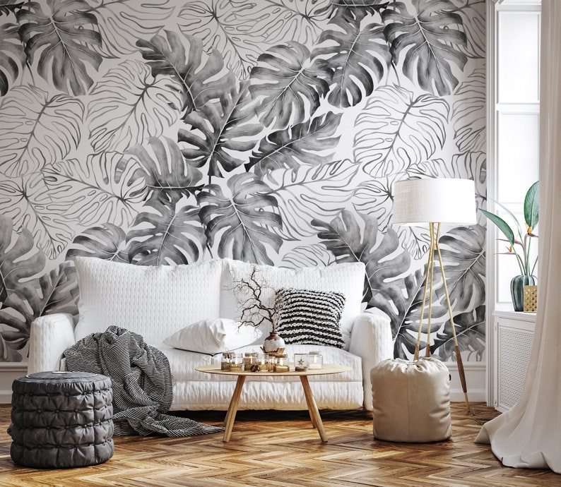 May include: A white sofa with a black and white throw blanket and a black and white patterned pillow sits in front of a wall with a black and white tropical leaf wallpaper. A small table with a vase of dried branches and a lamp with a white shade are in the room.