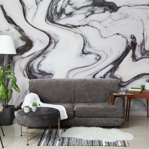 Peut inclure: Un canapé gris avec un jeté blanc est placé devant un mur avec un motif de marbre noir et blanc. Un petit pouf rond gris avec des pieds dorés est placé devant le canapé. Une petite table d'appoint en bois avec un verre et des livres est placée à droite du canapé.
