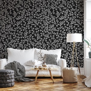 May include: A white couch with black and white throw pillows sits in front of a black wall with a white floral pattern. A gray throw blanket is draped over the couch. A gray ottoman sits in front of the couch. A wooden coffee table with a vase of branches and candles sits in front of the couch. A floor lamp with a white shade stands next to the couch. A white curtain hangs in the window behind the couch.