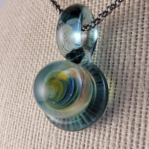May include: A teal glass pendant with a swirling pattern of yellow, blue, and green inside. The pendant is attached to a black chain.