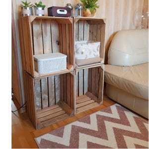 May include: A rustic storage unit constructed from wooden crates. The unit features four stacked crates, displaying plants, a white basket, and a small box. A patterned rug and a beige sofa are visible in the background.