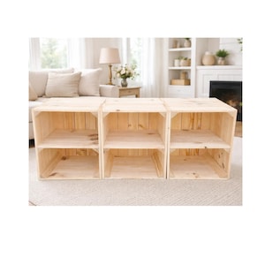 May include: Three unfinished wooden storage crates arranged side-by-side. Each crate has two shelves. The crates are made of light-coloured wood and are sitting on a light-coloured rug.