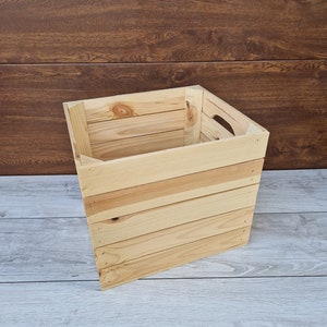 May include: A natural wood crate with a rectangular shape and a handle on the side. The crate is made of unfinished wood and has a rustic look.