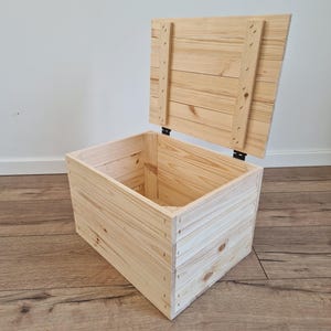 May include: A rectangular wooden crate with a hinged lid that is propped open. The box is constructed from light-coloured pine planks with visible wood grain and knots, featuring metal hinges connecting the lid to the main body of the container.