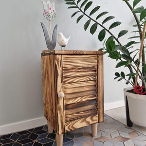 May include: A small, rustic wooden cabinet with a single louvered door and a light-coloured knob. The cabinet is stained a warm brown and sits on four short, light-coloured legs. A felt tulip and a white ceramic bird are on top.