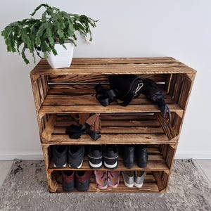 May include: A wooden shoe rack constructed from repurposed crates. The rack features three shelves, holding shoes, a purse, and an umbrella. A potted plant sits on top. The wood has a warm, brown finish.