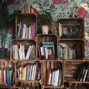 May include: A collection of wooden crates repurposed as bookshelves, filled with various books. The crates are arranged in a stacked configuration against a floral wallpaper background. A potted plant is visible on the top shelf.