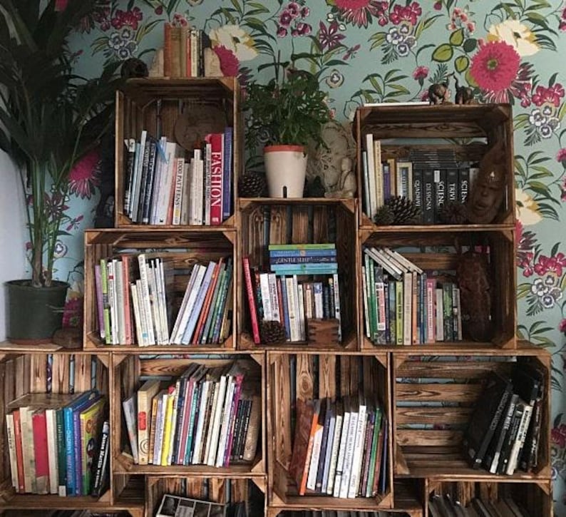 May include: A collection of wooden crates repurposed as bookshelves, filled with various books. The crates are arranged in a stacked configuration against a floral wallpaper background. A potted plant is visible on the top shelf.