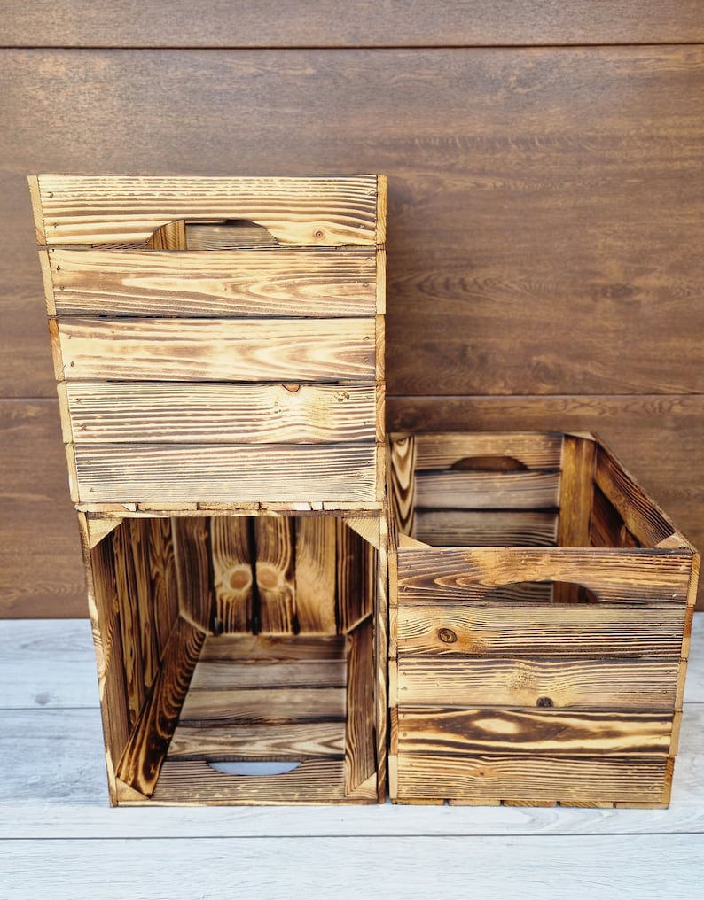 May include: Three wooden crates stacked on top of each other. The crates are made of natural wood with a rustic, weathered look. The crates are empty and have a rectangular shape.