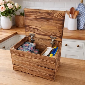 May include: A wooden storage box with a hinged lid, open to reveal contents. The box is made of light brown wood with visible grain. Inside are floral-patterned boxes, pens, and other small items. The box sits on a wooden countertop.