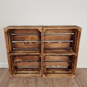 May include: Two rustic wooden crates, stained in a warm brown hue, are positioned side-by-side. Each crate features a slatted design with interior shelves, offering storage or display space. The wood grain is visible, adding to the natural aesthetic.