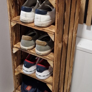May include: A rustic, wooden shoe rack with multiple shelves, holding various pairs of shoes. The rack is stained a warm brown and has a plant in a white pot on top. The shoes include sneakers and casual footwear.