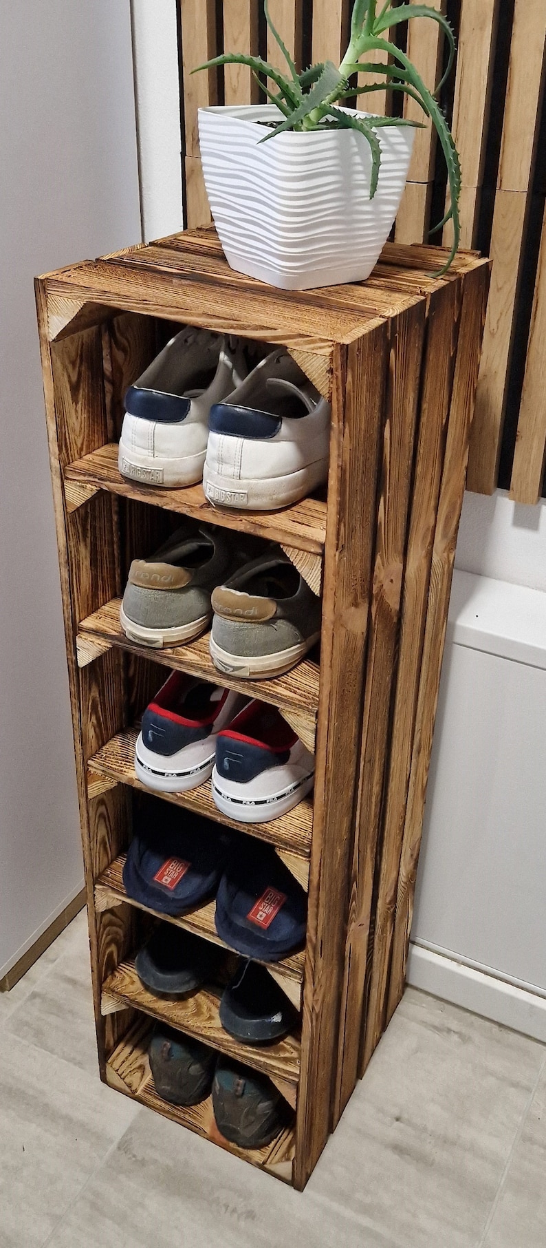 May include: A rustic, wooden shoe rack with multiple shelves, holding various pairs of shoes. The rack is stained a warm brown and has a plant in a white pot on top. The shoes include sneakers and casual footwear.
