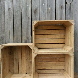 May include: Three unfinished wooden crates of varying sizes are stacked against a weathered wooden background. The crates are constructed with horizontal slats and corner supports, showcasing a rustic aesthetic. The wood has a natural, light brown color.