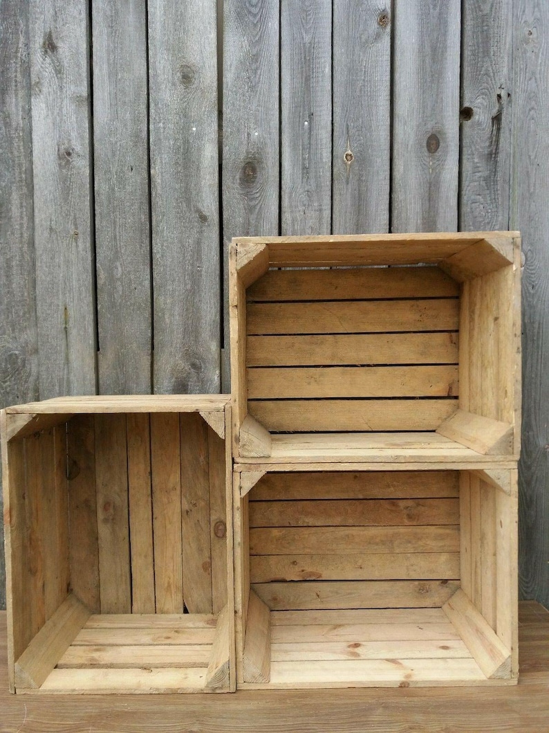May include: Three unfinished wooden crates of varying sizes are stacked against a weathered wooden background. The crates are constructed with horizontal slats and corner supports, showcasing a rustic aesthetic. The wood has a natural, light brown color.