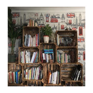 May include: A bookcase constructed from wooden crates, filled with books of different sizes and colours. The crates are stacked against a wallpaper backdrop featuring London landmarks and Union Jack flags. A potted plant is visible.