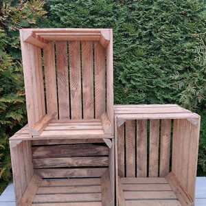 May include: Three wooden crates of varying sizes are stacked together. The crates are made of light brown wood and have a slatted design. The background is a dense green hedge.