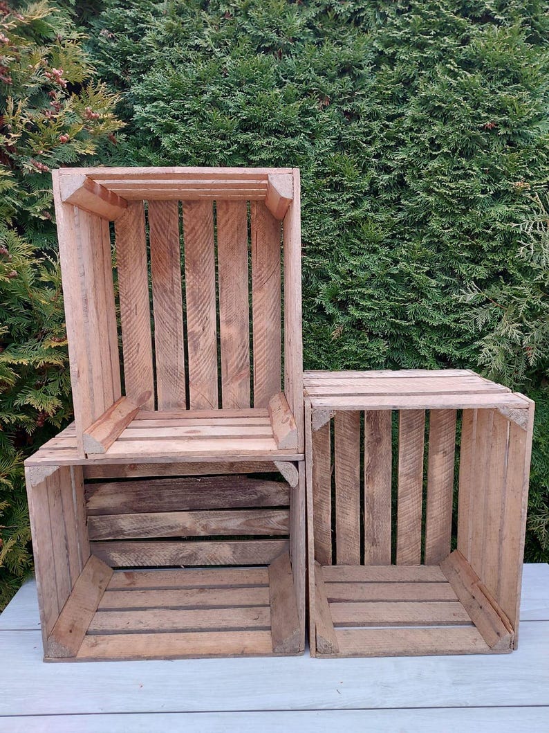 May include: Three wooden crates of varying sizes are stacked together. The crates are made of light brown wood and have a slatted design. The background is a dense green hedge.