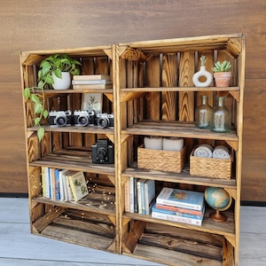 May include: Two wooden crate shelves, stained a warm brown, are filled with decorative items. The shelves hold books, cameras, plants, small baskets, glass bottles, and a globe. The shelves are arranged against a wooden wall.