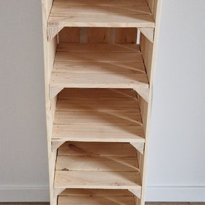 May include: A tall, unfinished wooden shelf unit with five shelves. The shelves are made of light-colored wood and are supported by a frame of the same material. The unit is standing on a wooden floor against a white wall.
