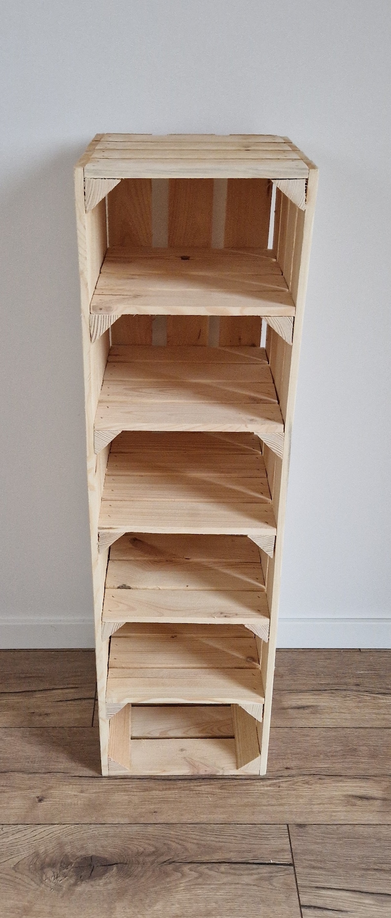 May include: A tall, unfinished wooden shelf unit with five shelves. The shelves are made of light-colored wood and are supported by a frame of the same material. The unit is standing on a wooden floor against a white wall.