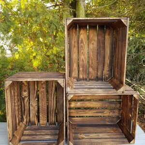May include: Three wooden crates with a dark brown finish are stacked against a backdrop of green foliage. The crates are constructed with vertical wooden slats and corner supports, showcasing a rustic aesthetic. The crates are empty.