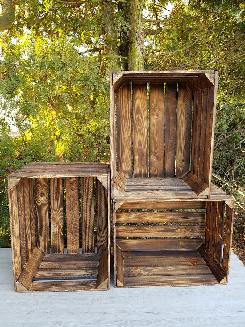 May include: Three wooden crates with a dark brown finish are stacked against a backdrop of green foliage. The crates are constructed with vertical wooden slats and corner supports, showcasing a rustic aesthetic. The crates are empty.