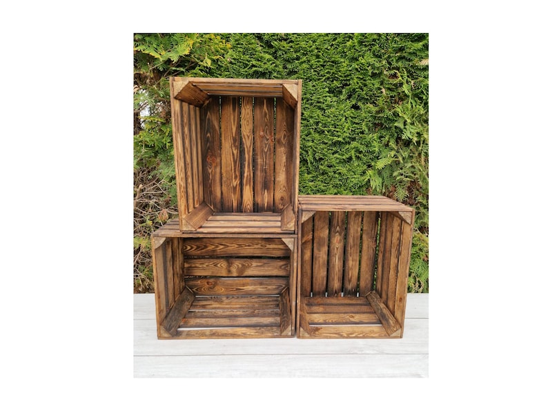 May include: Three rustic wooden crates, stained a rich brown, are stacked against a backdrop of green foliage. The crates are open-topped and constructed with visible wood grain, showcasing a natural, weathered aesthetic. Ideal for storage or display.