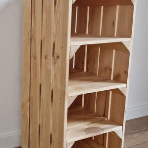 Puede incluir: Una unidad de almacenamiento alta y rectangular construida con listones de madera natural de color claro. La estructura cuenta con tres estantes abiertos con soportes de esquina reforzados, creando un dise&ntilde;o vertical tipo caja con veta de madera visible.