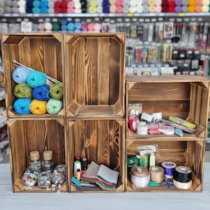 May include: Six wooden crates filled with craft supplies. The crates contain yarn, buttons, spools of ribbon, fabric squares, and other crafting materials. The crates are arranged in a display.