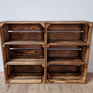 May include: Two rustic wooden crates joined together, forming a shelving unit. The shelves are made of dark brown, weathered wood with visible grain. Each crate has three shelves, perfect for storage or display. The unit is set against a white wall and wooden floor.