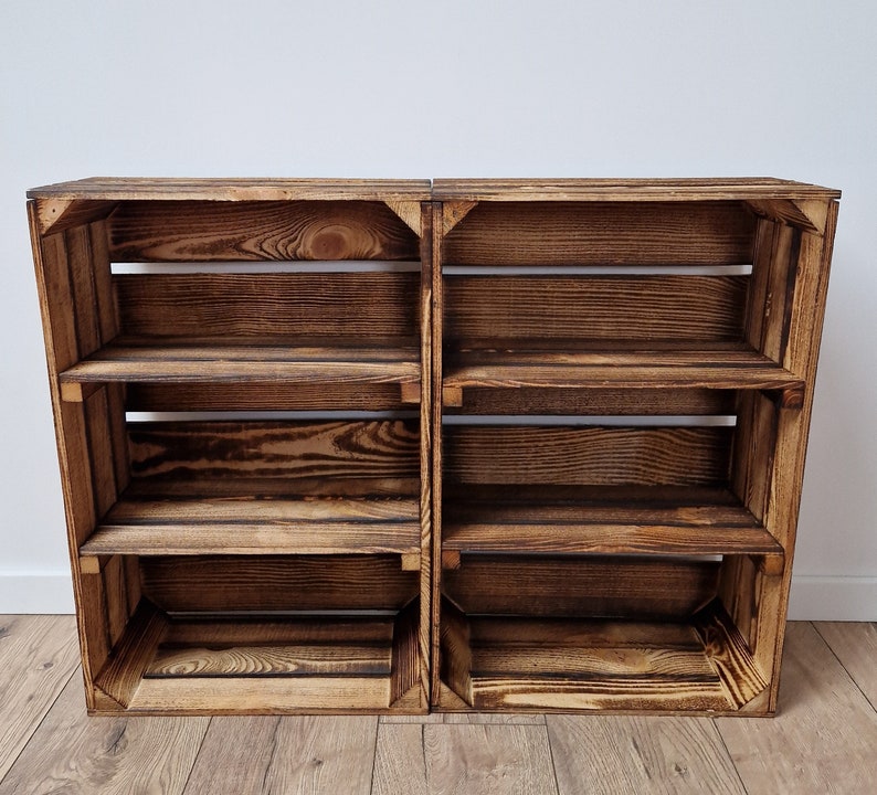 May include: Two rustic wooden crates joined together, forming a shelving unit. The shelves are made of dark brown, weathered wood with visible grain. Each crate has three shelves, perfect for storage or display. The unit is set against a white wall and wooden floor.