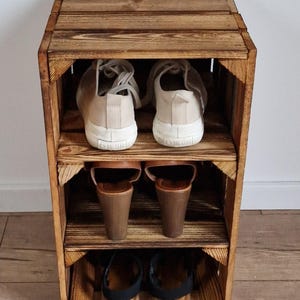 Puede incluir: Una unidad de almacenamiento de tres niveles construida con cajas de madera te&ntilde;idas de oscuro. El estante superior contiene un par de zapatillas beige, el estante central sandalias de cu&ntilde;a marrones y el inferior sandalias negras con tiras.