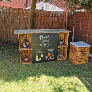 May include: A rustic outdoor bar made from wooden crates, featuring a chalkboard sign that reads "Beer and Wine." The bar has shelves displaying bottles and glasses. A matching crate with a gray cushion serves as a stool.