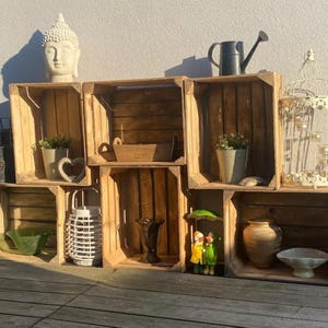 May include: A display of wooden crates arranged to create shelves, showcasing various decorative items. Items include a white Buddha head, potted plants, a watering can, a birdcage, a lantern, and figurines. The crates are made of weathered wood.