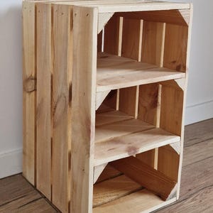 Puede incluir: Una estanter&iacute;a de caja de madera de color marr&oacute;n claro con listones verticales en los laterales y dos estantes interiores horizontales. La estructura est&aacute; hecha de madera de pino natural con vetas visibles y soportes de esquina reforzados.