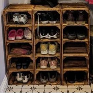 May include: Three wooden crate shoe racks, each with multiple shelves, are arranged side-by-side. Various shoes, including sneakers, sandals, and boots, are neatly organized on the shelves. The crates are made of light brown wood.