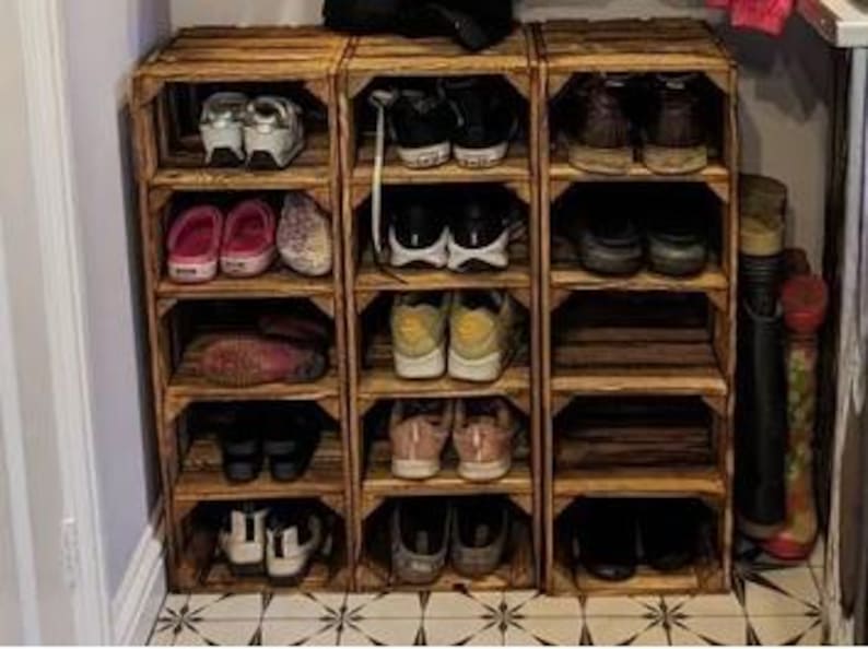 May include: Three wooden crate shoe racks, each with multiple shelves, are arranged side-by-side. Various shoes, including sneakers, sandals, and boots, are neatly organized on the shelves. The crates are made of light brown wood.
