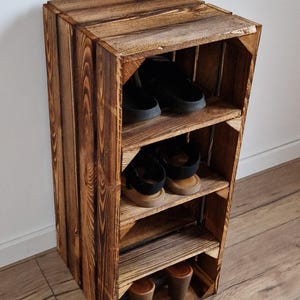 Puede incluir: Una unidad de almacenamiento vertical construida con cajas de madera te&ntilde;idas de oscuro con tres estantes horizontales. La unidad presenta patrones de veta de madera visibles y contiene tres pares de zapatos, incluyendo zuecos negros, sandalias y zapatos de tac&oacute;n marrones.