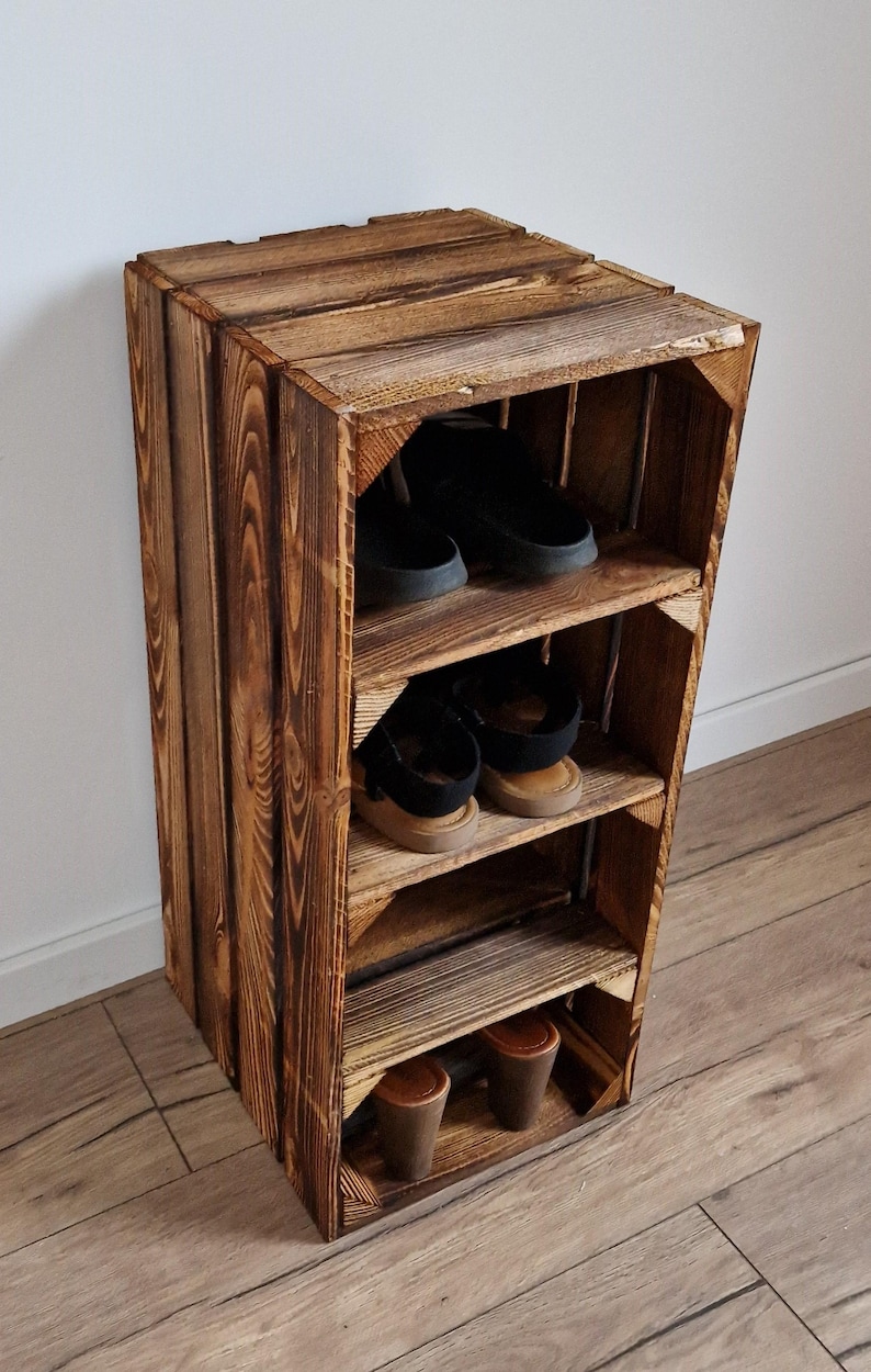 Puede incluir: Una unidad de almacenamiento vertical construida con cajas de madera te&ntilde;idas de oscuro con tres estantes horizontales. La unidad presenta patrones de veta de madera visibles y contiene tres pares de zapatos, incluyendo zuecos negros, sandalias y zapatos de tac&oacute;n marrones.