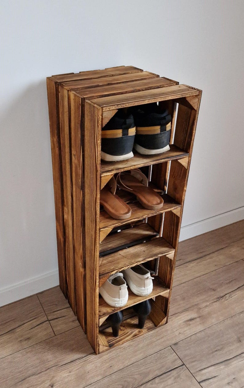 Puede incluir: Un zapatero vertical construido a partir de una caja de madera te&ntilde;ida con cuatro estantes horizontales. El estante contiene varios calzados, incluyendo zapatillas negras y amarillas, sandalias marrones, zapatillas beige y tacones negros.