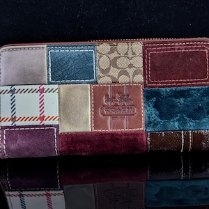 Może przedstawiać: Portfel typu patchwork z różnymi fakturami i kolorami, w tym zamszem, skórą i wzorzystymi tkaninami. Portfel posiada zapięcie na zamek błyskawiczny i pasek na nadgarstek. Logo Coach wytłoczone na skórzanej naszywce. Długość ok. 20 cm.
