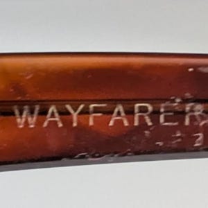 May include: Close-up of a brown Wayfarer eyeglass frame. The word "WAYFARER" is etched in silver lettering on the frame. The frame has a tortoiseshell pattern.