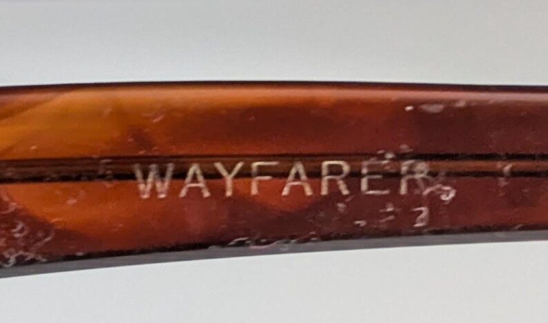 May include: Close-up of a brown Wayfarer eyeglass frame. The word "WAYFARER" is etched in silver lettering on the frame. The frame has a tortoiseshell pattern.